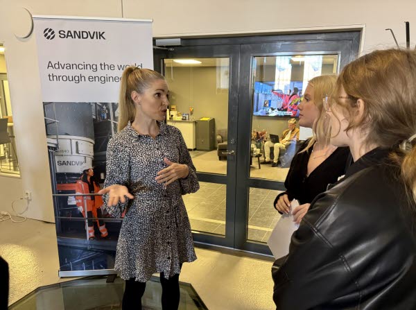 En kvinna står och pratar med två kvinnor i en lokal. I bakgrunden syns en roll-up med reklam för Sandvik. Foto. 