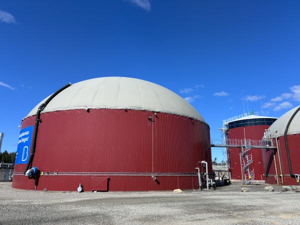 En stor rund och röd cistern som står på en grusplan. I kanten ser man en brygga som går över till en ny cistern. Foto. 