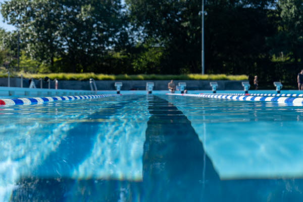 Stor pool omgiven av grönt gräs. Människor som både badar och ligger på gräset. Foto.