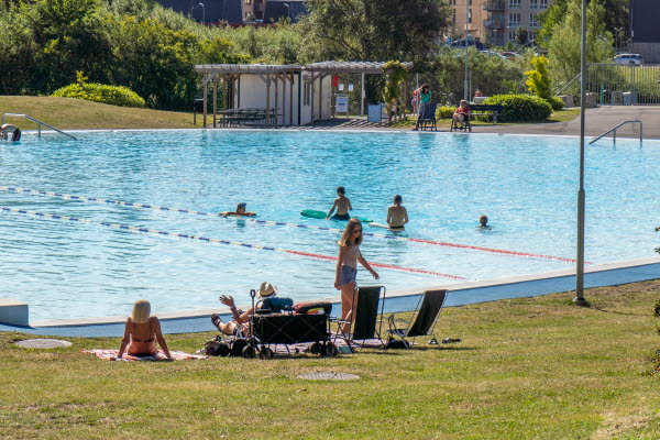 Ett fåtal personer i och kring en utomhusbassäng. Foto.