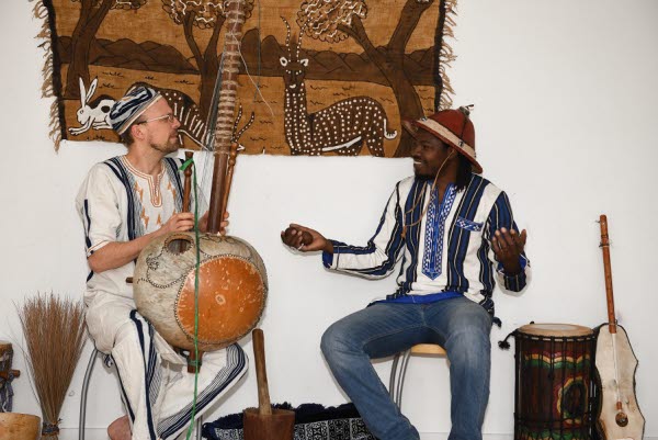 Två vuxna personer sitter ner pallar. Den ena med ett västafrikanskt instrument. Foto.