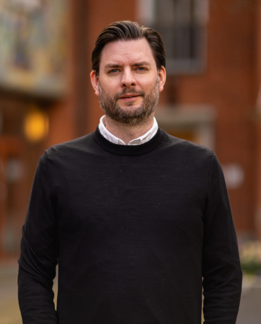 En portr&auml;ttbild av en man med kortklippt h&aring;r och sk&auml;gg, kl&auml;dd i l&aring;ng&auml;rmad tr&ouml;ja med skjorta under. Foto.