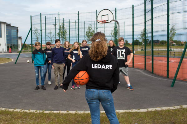 Ungdom står med en boll under armen vänd med ryggen emot kamera. På ryggen står "Ledare". Framför står en grupp ungdomar på en basketboll-plan. Foto.