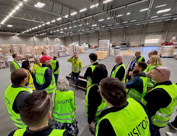 Ett femtontal personer i gula västar står i en ring i en stor lagerlokal och lyssnar på en person. Foto.