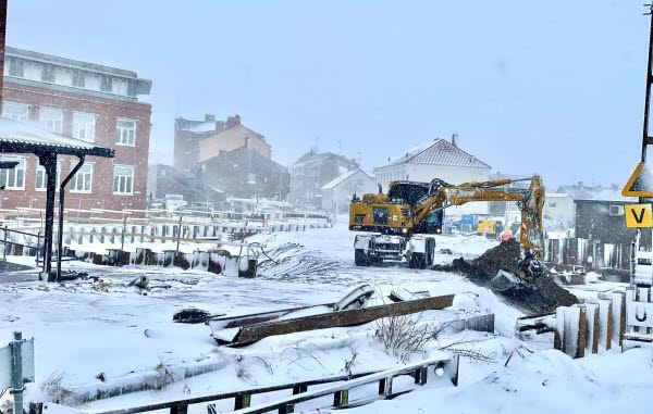 En gul grävmaskin gräver i ett snölandskap. I bakgrunden syns flera hus. Foto. 