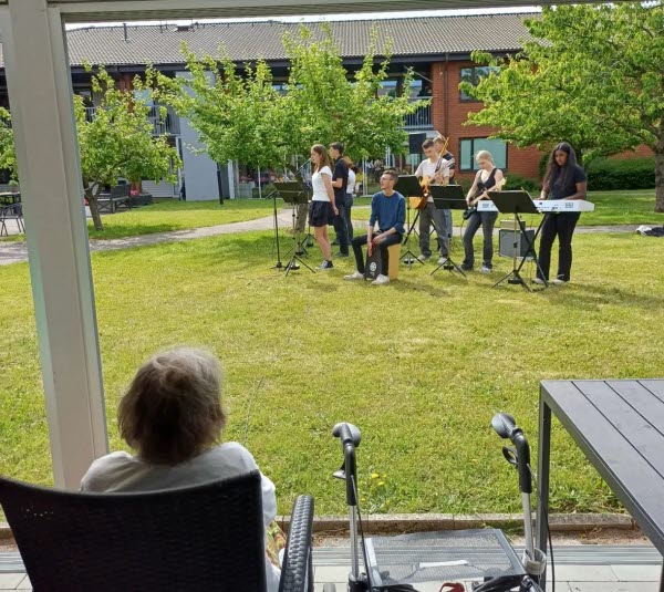 Ungdomar står på en gräsmatta och spelar musik. I förgrunden ser man en äldre person som sitter i en stol och tittar på. Foto. 