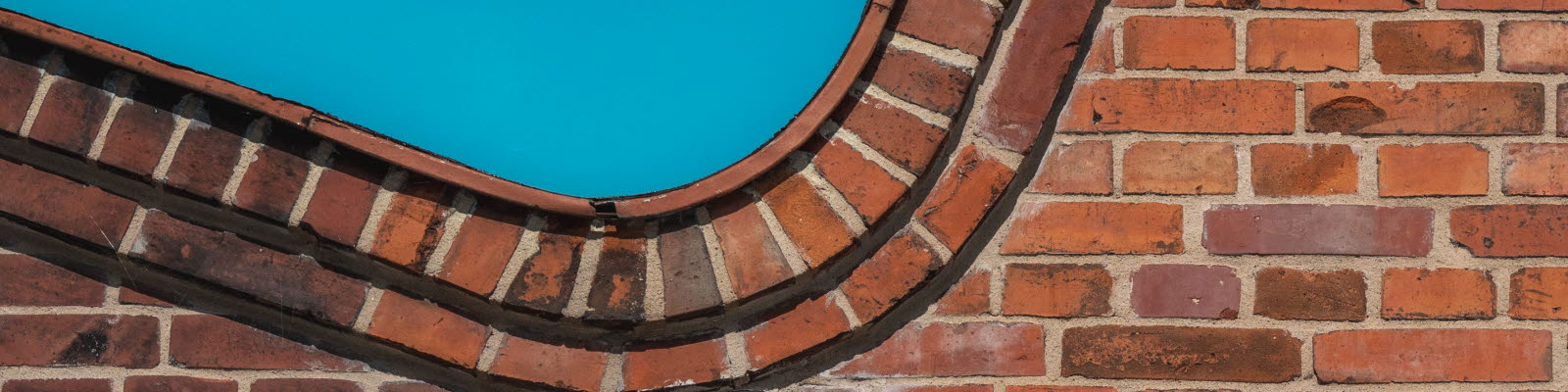 En gammal röd tegelbyggnad med stora fönster, utanför står ett grönt träd. Foto.