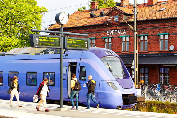 Ett blålila tåg framför en röd tegelbyggnad med texten "Svedala". Ett flertal människor går längs tåget. Foto.