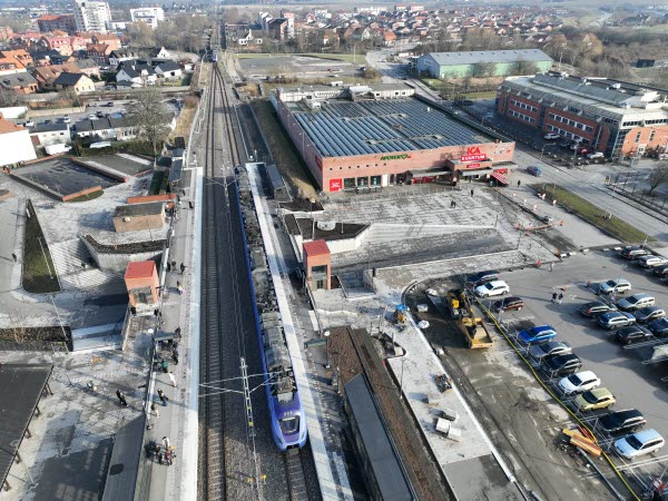 En drönarbild över stationsområdet i Svedala. Man ser två järnvägsspår, ett lila pågatåg, en bilparkering och en ICA-affär. I horisonten syns delar av Svedala. Foto.  