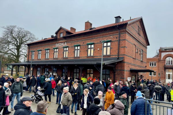 Folksamling framför stationshuset som är i rött tegel. Foto.