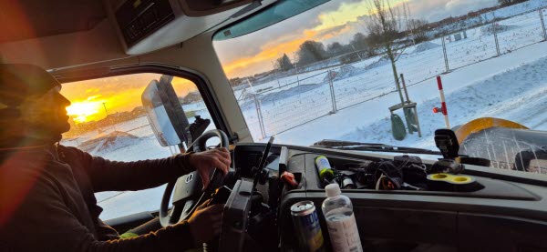 En man sitter bakom ratten i en lastbil. Det är snö ute och solen går upp. Foto. 