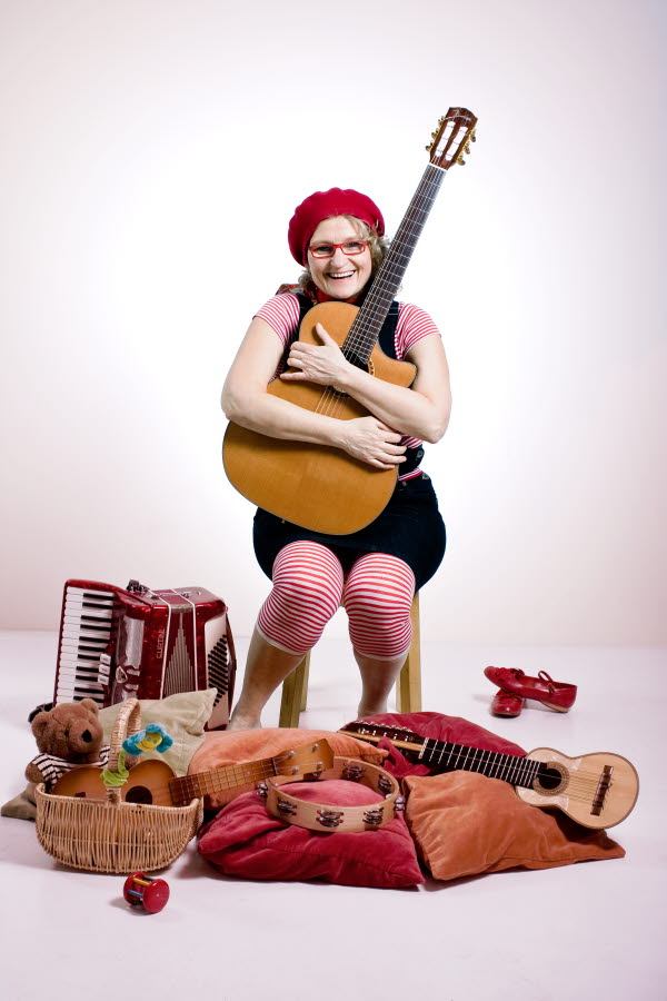 En person med röd basker och rödrandiga strumpor sitter med en gitarr i knäet. Foto.