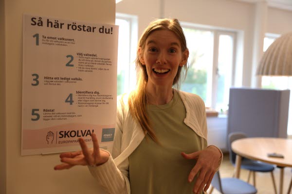 Lärare ler mot kameran och visar upp en affisch med instruktioner om hur du röstar i EU-valet. Foto.