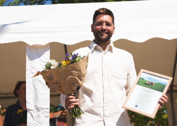 En man i vit skjorta står med blommor i handen och ler mot kameran. Foto.