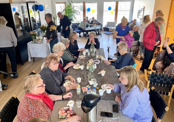 Åtta kvinnor sitter vid ett bord och fikar. I bakgrunden rör sig flera personer i lokalen. Foto.