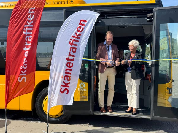Två personer står i dörröppningen på en gul buss och klipper ett bad. Framför syns röd och vita flaggor med texten Skånetrafiken. Foto. 