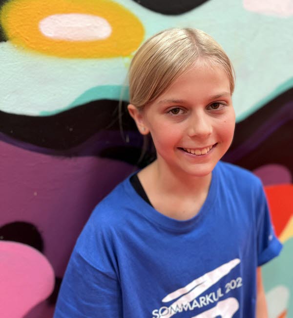 Flicka med ljust uppsatt hår och blå t-shirt med texten "Sommarkul 2023", står lutad mot en färgglad vägg. Foto.
