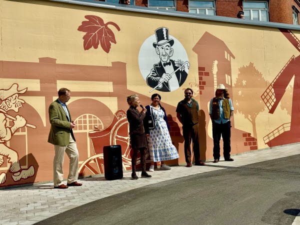 Fem personer står framför en målad mur och en av dem pratar i en mikrofon. Foto.