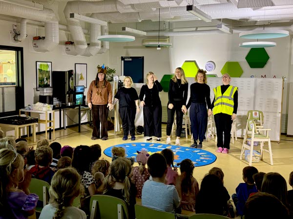 Sex personer håller varandra i handen  och står framför barn som sitter och tittar på golvet. Foto. 