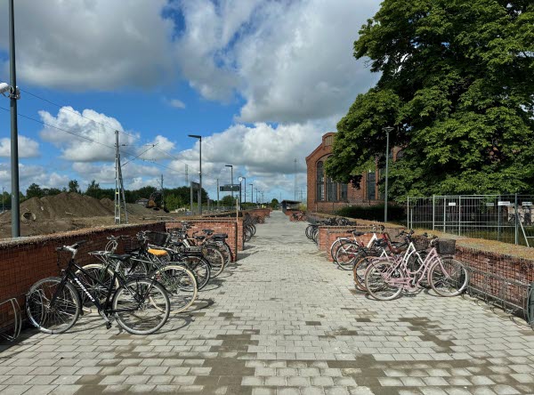 Cyklar står i ett cykelställ omgärdat av tegelmurar. I bakgrunden ser man delar av ett tegelhus. Foto. 